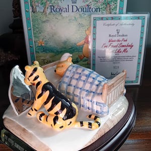 May include: A Royal Doulton figurine featuring Winnie-the-Pooh in bed and Tigger sitting nearby. The figurine is hand-decorated and includes a certificate of authenticity. The base reads "I've Found Somebody Just Like Me."