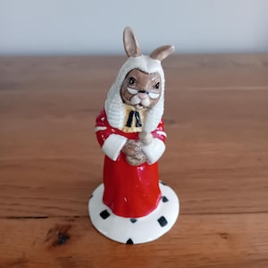 Puede incluir: Figura de cerámica de un conejo vestido con una túnica roja con ribetes blancos, una peluca blanca y gafas. El conejo sostiene un pergamino. La figura se apoya en una base blanca con detalles de diamantes negros.