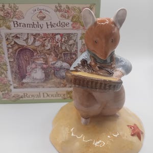 Può includere: Statuina in ceramica di un topo con un cestino, parte della collezione "Brambly Hedge" di Royal Doulton. Il topo è marrone e beige, indossa una camicia blu e pantaloni marroni. La statuina è su una base color sabbia con una foglia rossa.