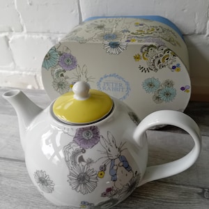 Puede incluir: Tetera de cerámica blanca con tapa amarilla y asa curva. La tetera está decorada con una ilustración de Peter Rabbit y diseños florales. Una caja a juego con el logotipo de Peter Rabbit está en el fondo.