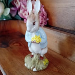 May include: A ceramic figurine of a rabbit standing on a textured green base. The rabbit has brown fur, wears a light blue jacket, and holds a small cluster of yellow flowers in its paws. The figurine features painted details and a glossy finish.