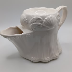 Op de afbeelding: Witte keramische scheerbeker met een decoratief ontwerp van schelpen en krulvormen. De beker heeft een ingebouwde zeepbak en een handvat.