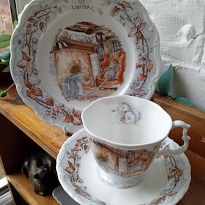 May include: A white ceramic plate, teacup, and saucer set. The plate has a scalloped edge and features a winter scene with a rabbit. The teacup and saucer have matching illustrations and floral accents.