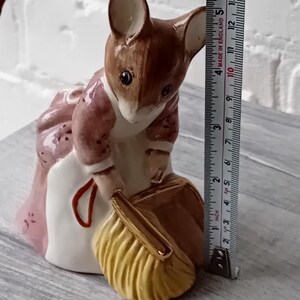 Puede incluir: Figura de cerámica de un ratón con un vestido rosa y un bolso amarillo. El ratón mide unos 10 cm de altura. La figura tiene detalles marrones y blancos. Fondo de pared de ladrillo blanco.