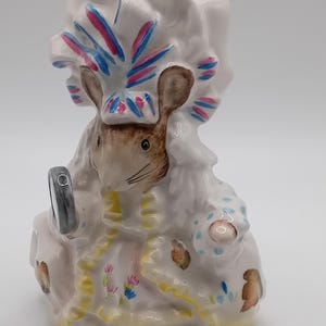 Puede incluir: Una figurita de cerámica blanca de un ratón con un gorro blanco con detalles en rosa y azul. El ratón sostiene un pequeño espejo y lleva un vestido blanco con detalles florales amarillos y rosas.