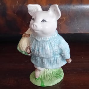 May include: A ceramic figurine of a pig standing on a green base. The pig is wearing a light blue textured jumper and carrying a yellow bundle under one arm. The figure features white skin, pointed ears, and detailed facial features in a glossy finish.