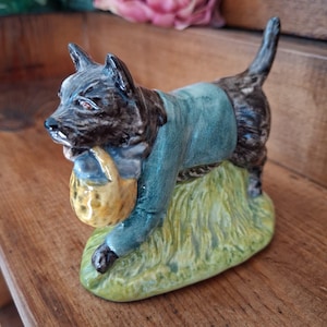May include: A ceramic figurine of a dark brown dog wearing a teal jacket and carrying a yellow basket in its mouth. The dog stands on a textured green base that resembles grass, with its tail pointed upward and its head turned to the side.