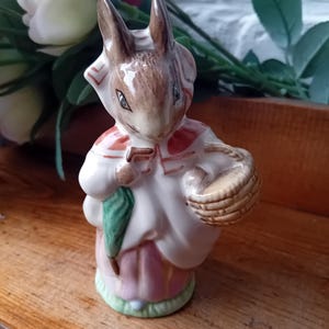 May include: A ceramic rabbit figurine wearing a white dress with pink and red details, carrying a green umbrella and a woven basket. The rabbit has brown fur and a white bonnet. The figurine stands on a green base.