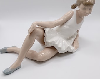 以下が含まれることがあります： 白いバレリーナの衣装を着た若い女の子の磁器製フィギュア。彼女は足を組んで座り、腕を膝の上に置いている。髪は2つの三つ編みに結ばれている。