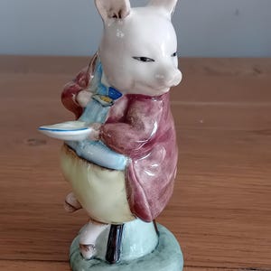 May include: A ceramic figurine of a pig wearing a maroon coat, blue shirt, and yellow pants. The pig is holding a small plate and is standing on a light blue base. The figurine has a whimsical, cartoonish design.