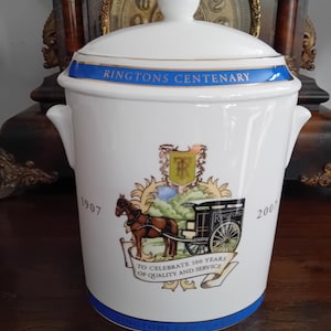 Może przedstawiać: Biały ceramiczny pojemnik z pokrywką i złotym uchwytem. Pojemnik posiada niebieski pasek z napisem RINGTONS CENTENARY, lata 1907 i 2007 oraz grafikę przedstawiającą powóz konny z napisem TO CELEBRATE 100 YEARS OF QUALITY AND SERVICE.