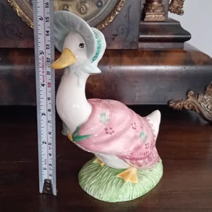 Puede incluir: Figura de cerámica de un ganso con gorro y vestido rosa con detalles florales, sobre una base verde. La figura mide aproximadamente 15 cm de altura.
