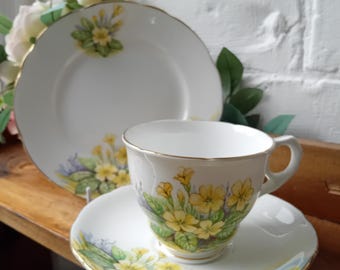 May include: A white teacup, saucer, and plate set with a floral design featuring yellow flowers and green leaves. The edges of the teacup, saucer, and plate are trimmed with gold. The teacup is on a saucer, and a plate is behind them.
