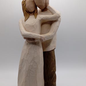 May include: A wooden sculpture of a couple embracing. The woman wears a cream-colored dress, and the man has brown pants and a white shirt. The sculpture has a carved texture and a neutral color palette, suggesting a focus on love and connection.