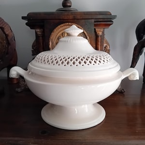 Puede incluir: Una sopera de cerámica blanca con tapa a juego que presenta un patrón decorativo de celosía calada alrededor del borde. La sopera tiene una base de pedestal y dos asas curvas en los lados, con un remate puntiagudo en el centro de la tapa.
