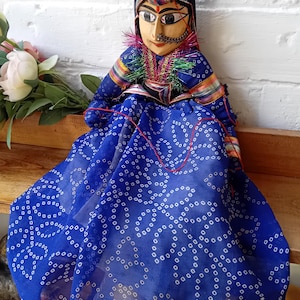 May include: A handcrafted wooden puppet doll with a painted face, wearing a blue patterned dress and a black head covering. The doll has a mustache and is adorned with colorful trim and jewelry. The doll is sitting on a wooden surface.