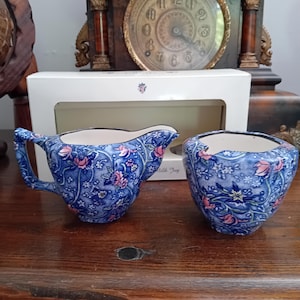 Könnte beinhalten: Ein Milchkännchen- und Zuckerdosen-Set aus Keramik mit blau-weißem Blumenmuster und rosa Akzenten. Die Teile haben einen dunklen Rand und stehen vor einer weißen Schachtel mit der Aufschrift Milk Jug sowie einer Uhr im Hintergrund.