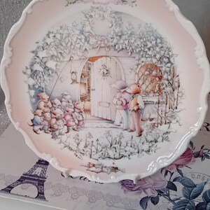 Op de afbeelding: Decoratieve plaat met een geschulpte rand en een roze rand. De plaat toont een winterse scène met dierenfiguren en de tekst "The Wind in the Willows". De plaat is waarschijnlijk van porselein.