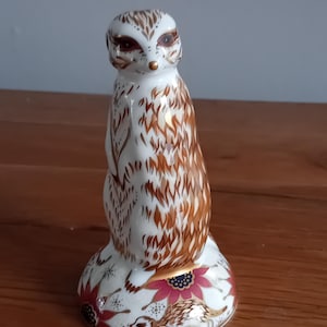 Puede incluir: Figura de suricato de cerámica. El suricato es blanco con detalles marrones y dorados. Tiene ojos marrones y una nariz dorada. La base presenta flores rosas y detalles dorados. Un artículo decorativo.