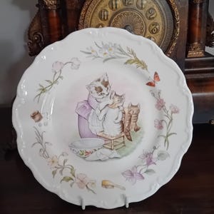 Peut inclure: Une assiette en céramique blanche à bord festonné présentant une illustration centrale d'un chat en robe violette lavant un chaton. La bordure est ornée de motifs floraux, d'un papillon, d'une bobine de fil et d'une petite brosse aux tons doux.