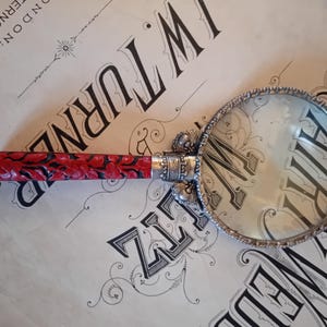 Könnte beinhalten: Eine Lupe mit einem silbernen Griff und einem rot-schwarzen gemusterten Griff. Der Griff ist mit silbernen Verzierungen versehen.