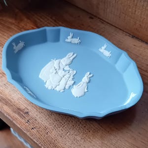 May include: A light blue oval decorative dish with a scalloped edge. The dish features white relief designs of rabbits and a group of rabbits with a basket. A charming piece for home decor.