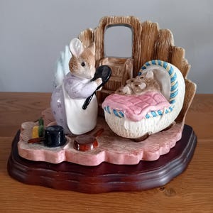 Puede incluir: Una figurita de porcelana de una madre conejo sosteniendo un espejo y mirando a sus conejitos bebés en una cuna. La figurita está colocada sobre una base de madera.