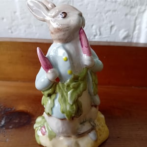 May include: A ceramic figurine of Peter Rabbit holding two carrots, one in each hand. He is wearing a blue jacket with yellow buttons and is standing on a green base.