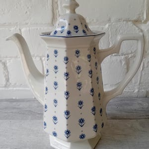 Könnte beinhalten: Eine weiße Kaffeekanne aus Keramik mit sechseckigem Körper und passendem Deckel. Die Oberfläche zeigt ein wiederkehrendes Muster aus kleinen blauen Blumen in vertikalen Reihen, akzentuiert durch winzige blaue Punkte zwischen den Motiven.