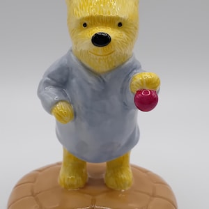 Op de afbeelding: Een geel keramiek beeldje van Winnie de Poeh, die een blauwe nachtjapon en een blauwe slaapmuts draagt. Hij houdt een rode bal in zijn rechterpoot en staat op een bruine, halvemaanvormige basis.