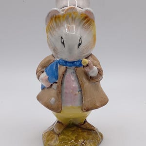 May include: A small ceramic figurine of a white guinea pig wearing a brown coat, blue scarf, yellow pants, and a white hat. The figurine is standing on a green base.