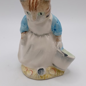 May include: A ceramic figurine of a cat wearing a blue jacket and white dress, holding a small bucket. The cat has brown and tan stripes on its head and is standing on a yellow and green base.