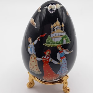 May include: A black, egg-shaped decorative item on a gold-coloured stand. The egg features a painted scene of three women in traditional dress dancing near a church. The egg is adorned with clouds.