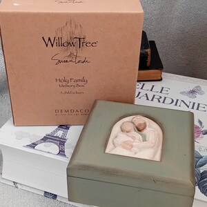 Könnte beinhalten: Eine Memory-Box aus salbeigrünem Holz mit einem geschnitzten Relief einer Familie. Die Box ist von Willow Tree und der Text "Holy Family Memory Box A child is born" ist auf der Box aufgedruckt. Hinter der Memory-Box befindet sich eine braune Box.