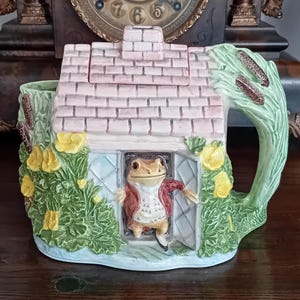 Puede incluir: Una caprichosa tetera de cerámica con forma de cabaña, con un personaje de rana con una chaqueta roja. La tetera tiene un tejado rosa, un asa verde y está adornada con flores amarillas y vegetación. Está sobre una superficie de madera.