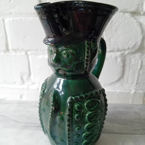 May include: A dark green glazed ceramic pitcher shaped like a figure wearing a military-style hat. The pitcher features molded details including a face, uniform buttons, and decorative trim. It has a curved handle on the side and a wide spout.