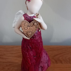 Puede incluir: Una figurita de un ángel con alas, vestida con un vestido rojo y sosteniendo un corazón de madera con la palabra "Love" en él. El ángel tiene una flor rosa en el pelo.