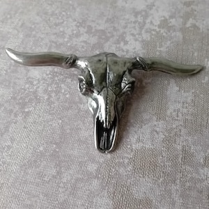Puede incluir: Una escultura plateada detallada de una calavera de toro con cuernos largos. La calavera tiene una superficie texturizada y se muestra sobre un fondo texturizado. Los cuernos se extienden hacia afuera, creando una estética rústica llamativa.