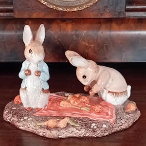 Puede incluir: Un conjunto de figuras de cerámica con dos conejos. Un conejo está de pie con una chaqueta azul, mientras que el otro está agachado sobre una manta roja con flores naranjas. La base es un óvalo marrón texturizado.