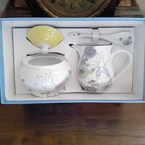 Puede incluir: Juego de té de cerámica blanca con diseño de conejos y flores, presentado en una caja azul. El juego incluye azucarero, jarrita para leche, cuchara y tapa amarilla. Diseño con detalles grises y morados.