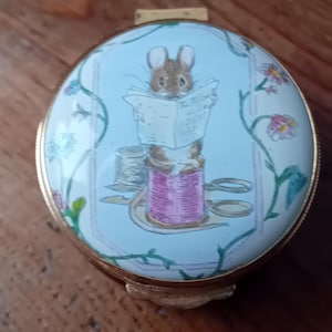 Puede incluir: Caja de baratijas ovalada con tapa azul claro. La tapa presenta un ratón de dibujos animados leyendo un periódico mientras está posado en una bobina de hilo rosa. La caja tiene un borde dorado y detalles florales.