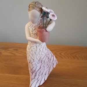 Puede incluir: Una figurita de cerámica blanca de una mujer con un vestido rosa y alas sosteniendo una pequeña maceta de terracota con flores rosas.