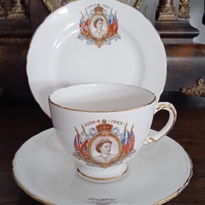 Op de afbeelding: Een witte porseleinen theekop en schotel set met een herdenkingsontwerp voor de kroning op 2 juni 1953, inclusief een portret van koningin Elizabeth II, gouden randen en kleurrijke vlaggen. Een bijpassend wit porseleinen bord staat erachter.