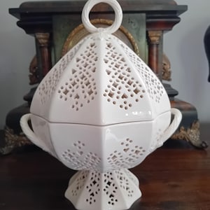 May include: White ceramic lidded serving dish with detailed cut-out patterns. The dish features a circular handle and two side handles. It stands on a pedestal base, displaying a classic design. The background includes a dark wooden surface and a vintage clock.