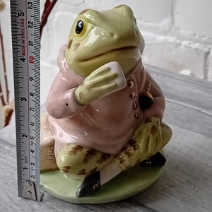 Puede incluir: Una figura de rana de cerámica, de aproximadamente 13 cm de altura, que lleva un abrigo rosa y sostiene un objeto blanco. La rana tiene patas verdes y marrones estampadas y está sentada sobre una base verde. Zapatos negros completan el atuendo.