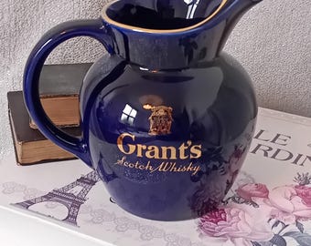 Puede incluir: Una jarra de cerámica azul con un borde dorado y un logotipo dorado de Grant's Scotch Whisky. La jarra está sobre un libro con un diseño floral.