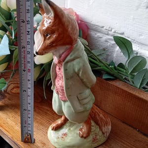 Puede incluir: Figura de cerámica de un zorro vestido con un abrigo verde y un chaleco rosa, de pie sobre una base verde. La figura mide aproximadamente 15 cm de altura. El zorro tiene pelaje marrón y está posando con las manos a la espalda. La figura está sobre una superficie de madera.