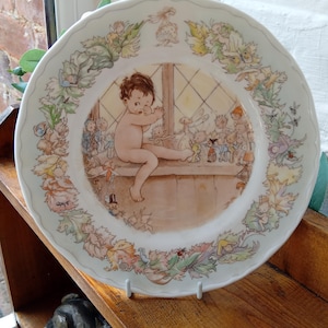 May include: A decorative porcelain plate with a charming illustration. The central image shows a nude child seated on a windowsill, surrounded by small figures. The border features a floral and fairy design, with a scalloped edge.