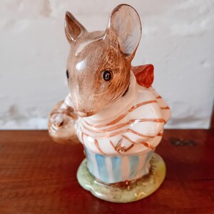 May include: A ceramic figurine of a brown mouse wearing a blue and white striped shirt and carrying a brown bag. The mouse has a red bow tie and is standing on a green base.