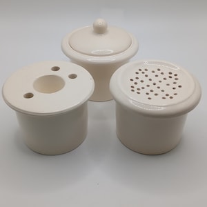 May include: Three white ceramic containers. One container has a lid, one container has a round opening with three holes, and one container has a lid with many small holes.
